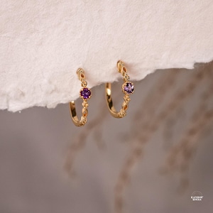 May include: A pair of gold-coloured hoop earrings, each with a small, round, purple gemstone. The earrings have a decorative, textured design along the hoop. The earrings are displayed against a white, textured background.