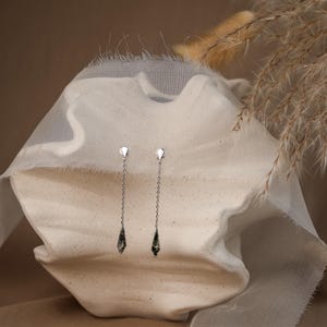 Puede incluir: Un par de pendientes colgantes plateados. Cada pendiente presenta una pequeña parte superior geométrica y una fina cadena que conduce a una gema facetada en forma de lágrima. Las gemas tienen un patrón verde oscuro y gris. Los pendientes se exhiben sobre una superficie blanca texturizada.