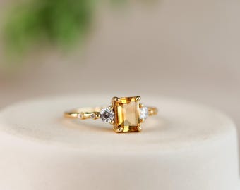 Puede incluir: Un anillo de oro con una piedra central de citrino amarillo de corte esmeralda rectangular, flanqueada por dos piedras blancas redondas más pequeñas. La banda es fina y de oro, con pequeñas piedras de acento a lo largo del aro.