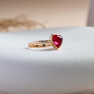 May include: A gold ring featuring a prominent heart-shaped red gemstone set in a bezel setting. The band is crafted from polished gold and includes a single small, round, clear accent stone embedded directly into the shank near the heart setting.