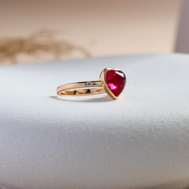 May include: A gold ring featuring a prominent heart-shaped red gemstone set in a bezel setting. The band is crafted from polished gold and includes a single small, round, clear accent stone embedded directly into the shank near the heart setting.