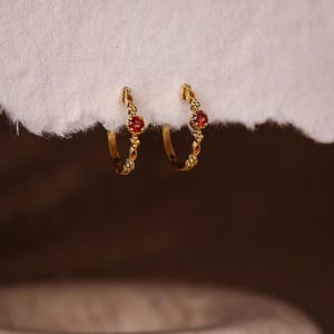 Könnte beinhalten: Ein Paar goldfarbene Creolen mit einem kleinen roten Edelstein in der Mitte. Die Ohrringe haben ein dekoratives, strukturiertes Design entlang des Reifens. Die Ohrringe werden auf einer weißen, strukturierten Oberfläche präsentiert.