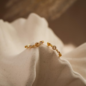 May include: Two delicate gold rings with intricate designs and small gemstones. The rings are displayed on a textured, off-white surface, with a soft, blurred background. The rings have a vintage or antique aesthetic.