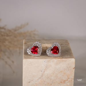 May include: A pair of heart-shaped stud earrings featuring a central red gemstone surrounded by a halo of small clear stones set in a silver-toned metal. The earrings are displayed side-by-side, showcasing the faceted red heart design.