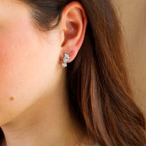 May include: A close-up of an ear with a silver stud earring. The earring has three light blue, irregular-shaped stones. The person has long, brown hair and is wearing a light green top.