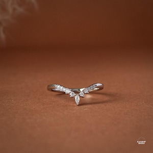 May include: A silver-toned V-shaped ring featuring a central marquise-cut stone flanked by several smaller round-cut stones along the band. The metal band has a polished finish and follows a chevron contour, creating a pointed design.