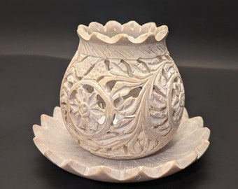 May include: A white stone candle holder with intricate floral carvings. The holder has a round base and a scalloped rim. It sits on a matching white stone plate.
