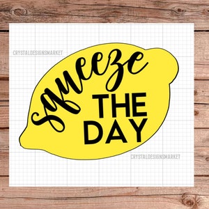 以下が含まれることがあります： レモンの形をした黄色のデザインで、黒い文字で「Squeeze the Day」と書かれています。
