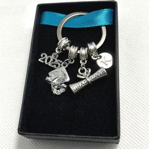 May include: A silver graduation keychain with a graduation cap, diploma, and the year 2023 charm. The keychain is in a black box with a blue ribbon.
