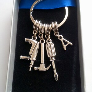 Puede incluir: Llavero plateado con dijes en forma de herramientas, incluyendo un taladro, un martillo y un destornillador, adem&aacute;s de una letra "A". El llavero se presenta en una caja de regalo con una cinta azul, sobre un fondo de terciopelo negro.