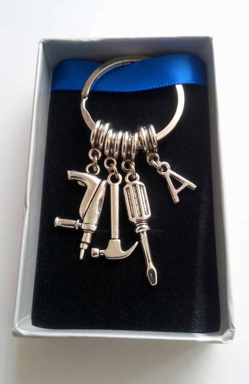 Puede incluir: Llavero plateado con dijes en forma de herramientas, incluyendo un taladro, un martillo y un destornillador, adem&aacute;s de una letra "A". El llavero se presenta en una caja de regalo con una cinta azul, sobre un fondo de terciopelo negro.