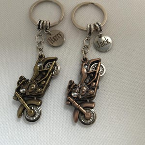 May include: Two motorcycle keyrings with silver key rings and small round charms. One charm reads "Harry" and the other reads "Jack". The motorcycle charms are bronze-coloured with detailed features.