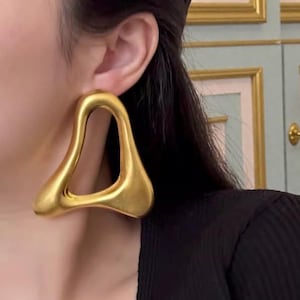 May include: Gold-toned, abstract-shaped earrings. The earrings have a smooth, matte finish and a unique, open design. The earrings are a statement piece, adding a touch of modern art to any outfit. The earrings are a bold accessory.