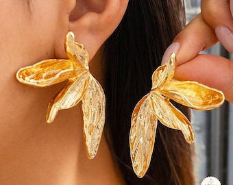May include: A pair of gold-colored, leaf-shaped earrings. The earrings have a textured surface and a three-dimensional design, with multiple petals extending outwards. The earrings are shown on a person's ear and held by a hand.