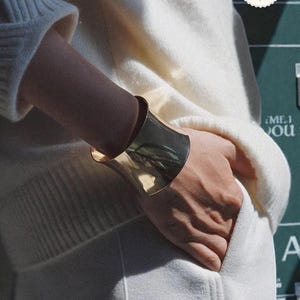 May include: A wide, polished gold-tone cuff bracelet. The bracelet is worn on a wrist, with the hand resting in a pocket of a white sweater. The bracelet has a smooth, reflective surface.