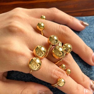 Puede incluir: Un conjunto de anillos dorados con grandes acentos redondos en forma de bola. Los anillos se llevan en varios dedos.