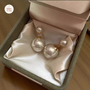 May include: A pair of pearl earrings with a gold post and backing, presented in a velvet-lined box. The earrings feature a large, round pearl and a smaller pearl on the post. The box is a muted green color, and the lining is a satin-like fabric.