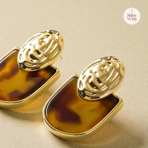 May include: Gold-coloured earrings with a tortoiseshell pattern. Each earring has a textured, oval top and a rectangular base with a glossy, brown and amber design. The earrings have a post-back fastening.
