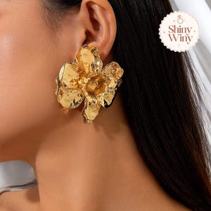 Könnte beinhalten: Goldfarbene Ohrringe in Blumenform. Die Ohrringe sind groß und haben eine strukturierte Oberfläche.