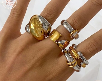 Könnte beinhalten: Eine Hand trägt einen Satz von fünf Ringen. Die Ringe sind aus Silber und Gold gefertigt und haben verschiedene Designs. Die Ringe sind übereinander auf dem Mittelfinger gestapelt.
