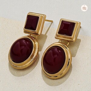 May include: A pair of gold-toned earrings with a square top and an oval bottom, each featuring a deep burgundy-colored stone. The earrings have a post-back closure and a polished finish, suitable for everyday wear or special occasions.