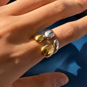 Könnte beinhalten: Ein goldener und silberner Ring mit einem einzigartigen, abstrakten Design. Der Ring hat zwei runde, glatte Kugeln, die durch ein gebogenes Band verbunden sind.