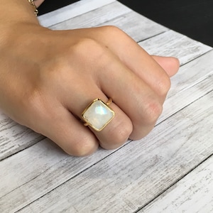 May include: A gold ring with a square-shaped moonstone gemstone. The ring is on a finger.