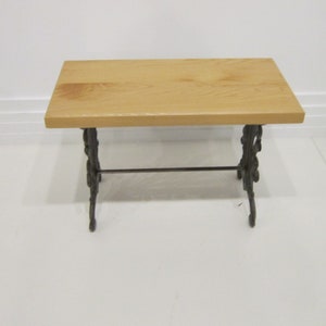 May include: A small wooden table with a natural wood finish and a black metal base. The table has a simple design with a rectangular top and a single metal bar connecting the legs.