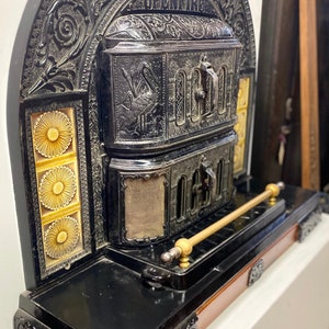May include: An antique open fireplace made of black cast iron with intricate detailing and a gold-coloured metal handle. The fireplace features a decorative tile panel with a floral design.