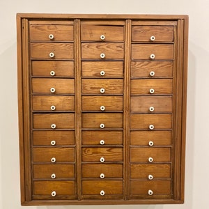 Puede incluir: Un armario de madera con 24 cajones, cada uno con una perilla blanca. El armario está hecho de madera marrón claro y tiene un diseño simple.