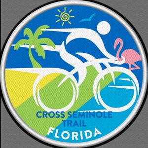 May include: A white bicycle with a person riding it, a palm tree, a sun, a pink flamingo, and the text "CROSS SEMINOLE TRAIL FLORIDA" on a blue, green, and yellow background.
