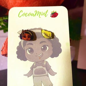 May include: A pair of earrings featuring a cartoon character with brown skin and a green leaf. The earrings are on a card with the text "CocoaMint" and "@CocoaMintLofi".