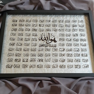 Puede incluir: Una pieza de arte de pared enmarcada en negro con fondo plateado. El arte presenta una gran inscripción árabe en negro, con la palabra "Allah"  desplegada prominentemente en el centro.