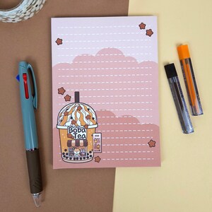 May include: A notepad featuring a pink cloud design and a Boba Tea illustration. The notepad has lined pages and star accents. A teal pen and two mechanical pencils are also present.