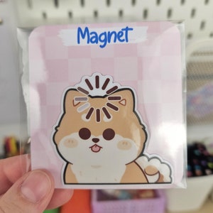 Puede incluir: Un imán con un diseño de perro Shiba Inu de dibujos animados. El imán está empaquetado en una funda transparente y tiene un fondo rosa con un patrón de cuadros. La palabra "Magnet" está escrita en azul.