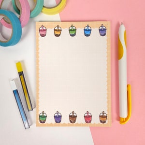 Könnte beinhalten: Ein Notizblock mit gewelltem Rand und Gittermuster, verziert mit bunten Bubble-Tea-Illustrationen. Ein weißer und gelber Stift, ein blauer und gelber Druckbleistift und bunte Washi-Tape-Rollen sind um den Notizblock auf einer weißen und rosa Oberfläche angeordnet.