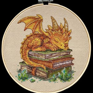 May include: Cross-stitch embroidery of a golden dragon resting on a stack of books. The dragon has detailed scales and wings, with green crystals at the base. The embroidery is set in a round wooden hoop.