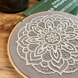 May include: A detailed embroidery hoop featuring a cream-colored floral design on a gray fabric. The design is centered and symmetrical, with a wooden hoop holding the fabric taut. The embroidery kit is visible in the background.