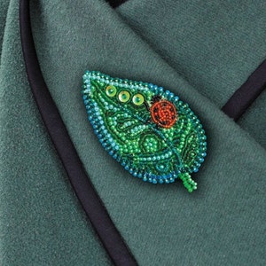 May include: A green leaf-shaped brooch with a red and black ladybug design, made of beads. The brooch is on a green fabric background.