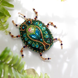 Può includere: Una spilla a forma di scarabeo verde con dettagli dorati e blu. La spilla è realizzata con perline e ha un design dettagliato.