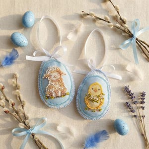 May include: Two embroidered Easter egg ornaments with a lamb and a chick, on a light blue background. The eggs have white ribbon hangers. Scattered around are blue speckled eggs, feathers, and pussy willow branches tied with blue ribbons.