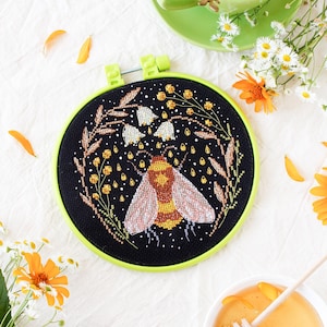 Może przedstawiać: Czarne tamborek do haftu z zieloną ramką, przedstawiające haft krzyżykowy pszczoły z żółto-brązowymi paskami, otoczonej białymi kwiatami i żółtymi kroplami deszczu na czarnym tle.