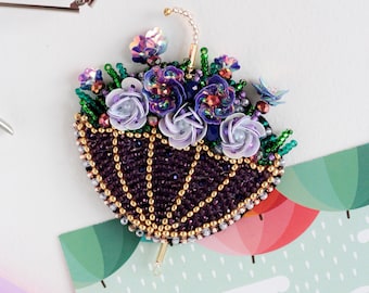 May include: A beaded brooch in the shape of an umbrella with purple, green, and gold beads. The umbrella is decorated with white and purple flowers and iridescent sequins.