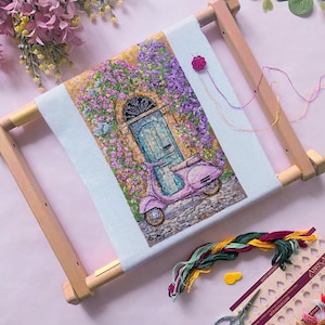 Puede incluir: Bordado de punto de cruz de una vespa rosa frente a una puerta azul, rodeada de flores moradas. La obra está montada en un marco de madera. También se ven hilos de bordar, tijeras y una aguja.