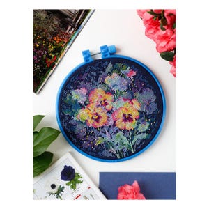 May include: A circular blue embroidery hoop containing a cross-stitch pattern of vibrant yellow, orange, and purple pansy flowers set against a dark navy blue background. The needlework features intricate thread details and colorful floral motifs.