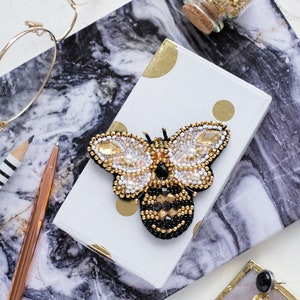 May include: A beaded bee brooch with black, white, and gold accents. The brooch is on a white box with gold polka dots.