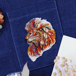 May include: A beaded lion head brooch with a detailed design in shades of orange, red, brown, and gold. The brooch is set against a dark blue fabric, showcasing the intricate beadwork and craftsmanship. The lion's mane is the focal point.