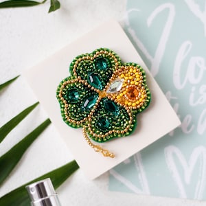 May include: A green and yellow beaded clover brooch with a gold chain attached. The brooch is on a white surface with a green background.