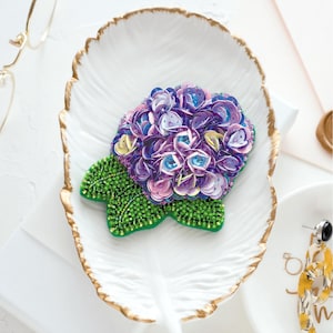 May include: A handcrafted brooch shaped like a hydrangea flower, featuring layered purple and blue sequins for the petals and small green seed beads forming the leaves, resting on a white leaf-shaped ceramic dish with a gold-painted rim.