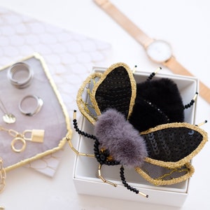 May include: A black and gold bee brooch with black beads and a fuzzy gray body. The brooch is in a white box with a white background.